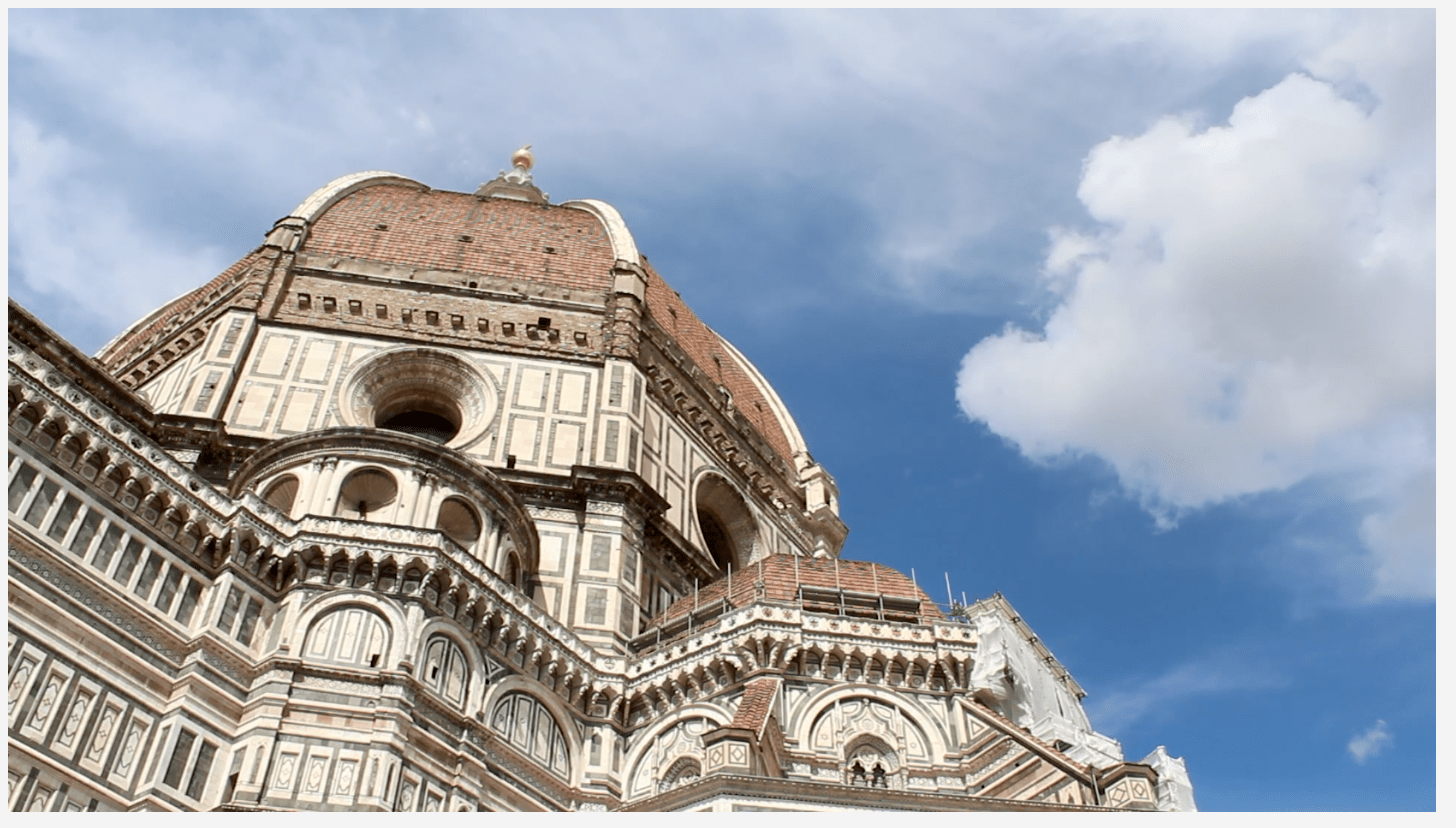 Upward shot of a cathedral dome in Italy. Sky is a beautiful blue with puffy, white clouds. Dome is a mix of gorgeous, architectural details in orange, brown and white.
