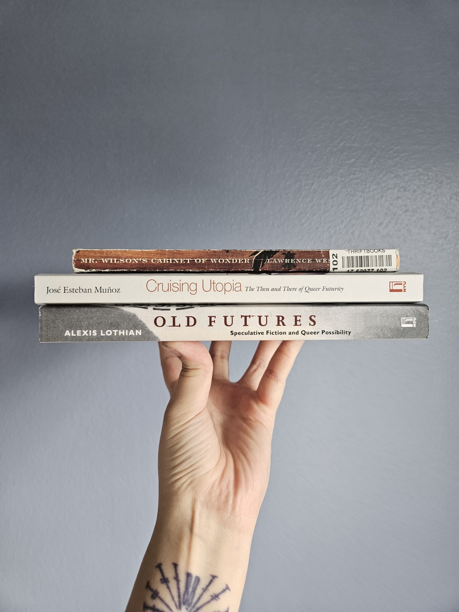 Hand holding up a stack of three books: Mr. Wilson's Cabinet of Wonder, Cruising Utopia and Old Futures.