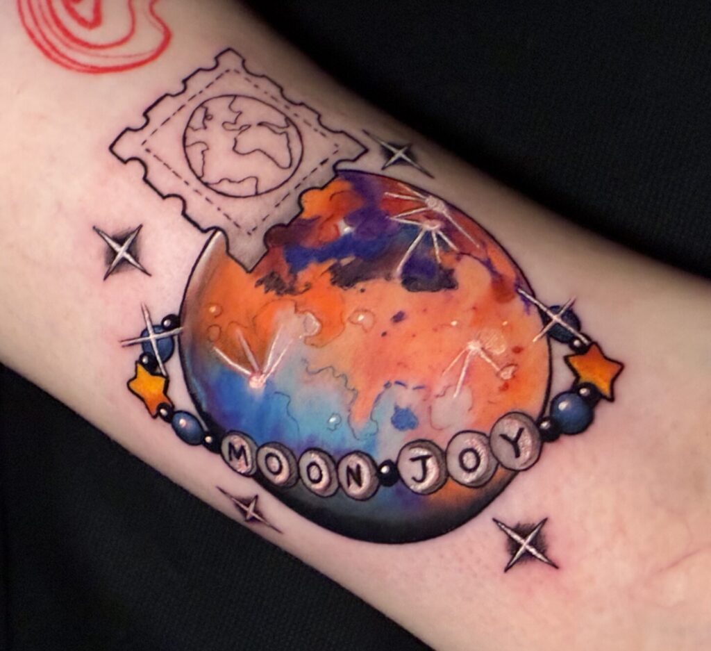 Colorful tattoo on white skin. Moon is encompassed by beaded bracelet with the words MOON JOY. A postage stamp depicts Earth. Four stars surround the Moon.