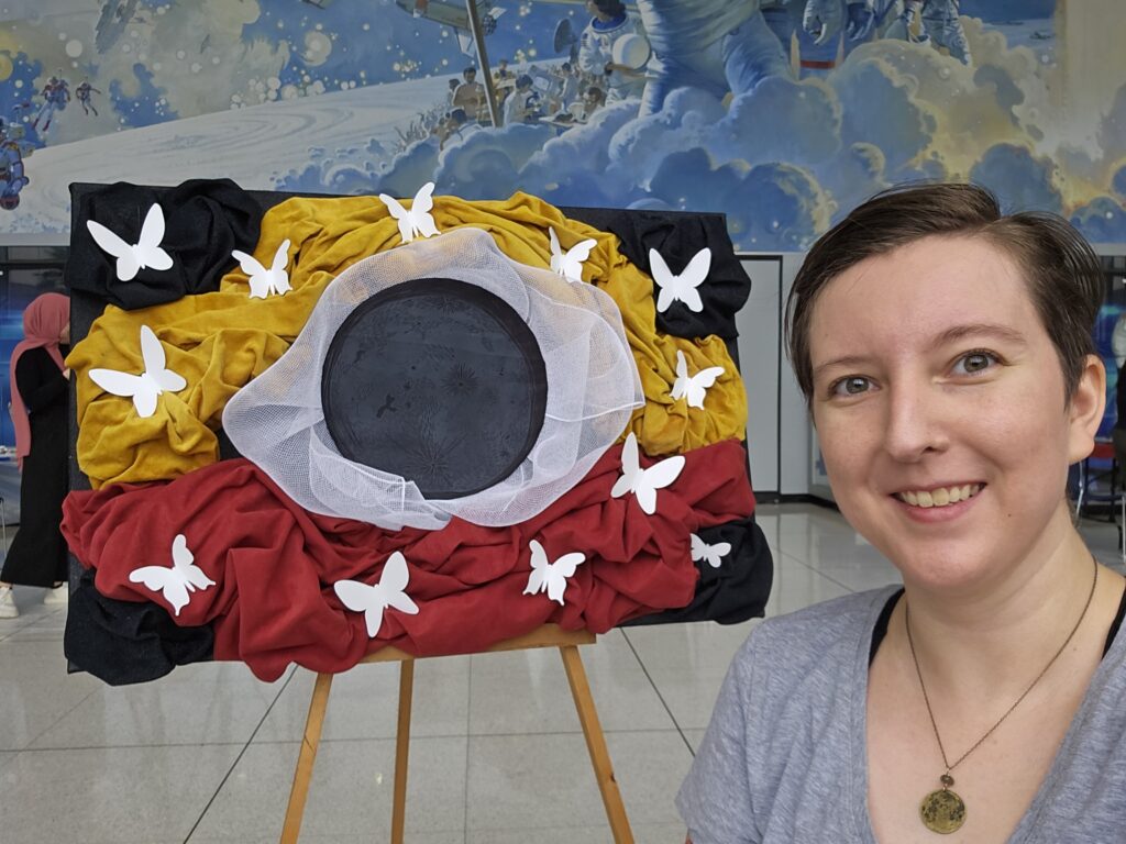 Artist with fabric sculpture. Moon print in middle on black fabric with white, yellow and red fabric bunched around. White butterflies fly outward.